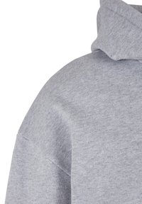 Détail d'un tissu gris clair de sweat à capuche montrant la couture d'épaule et la capuche avec un matériau tricoté doux et texturé sur fond blanc.