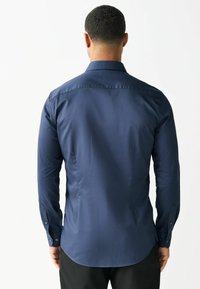 Man die van achteren is gezien, gekleed in een nauwsluitend, donkerblauw overhemd met lange mouwen en met knopen gesloten manchetten, en zwarte broek, tegen een witte achtergrond.