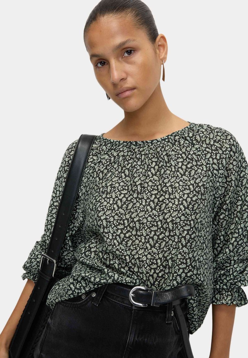 Black floral blouse with white patterns, round neckline, and gathered detailing. Paired with black jeans and a wide black belt.