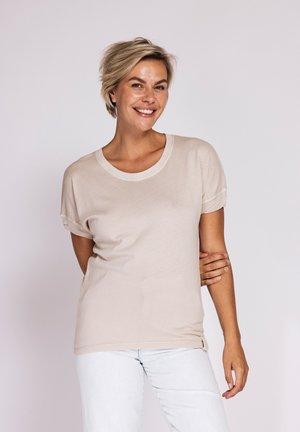 Smiling woman with short blonde hair wearing a beige T-shirt and white pants stands against a plain light background, one hand behind back.