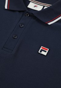 Marinblå pikétröja med texturerad tyg, tre knappar och en liten broderad logotyp i rött, vitt och blått på vänster bröst.