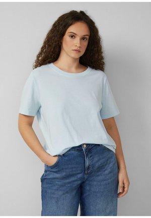 Frau mit lockigem Haar, die ein hellblaues, kurzärmeliges T-Shirt und blaue Jeans trägt, steht mit einer Hand in der Tasche vor einem neutralen Hintergrund.