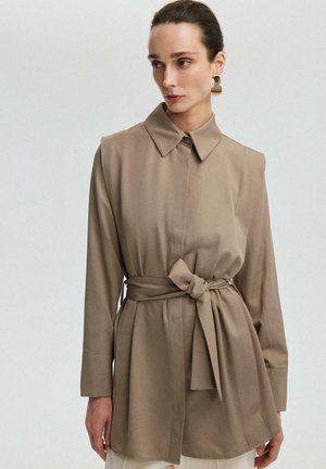 Sötét hajú nő bézs színű, öves, hosszú ujjú ingben és nagy, arany geometriai fülbevalókkal, balra néz egy egyszínű háttér előtt.