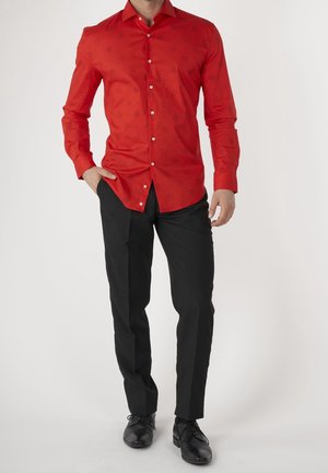 Mand iført en rød langærmet skjorte med subtile mønstre, sorte pæne bukser og sorte pæne sko, stående med den ene hånd i lommen.