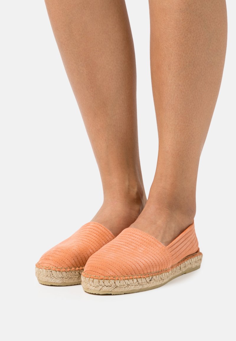 Pari di piedi che indossano scarpe espadrillas slip-on a coste color salmone con suole in juta intrecciata su uno sfondo bianco.