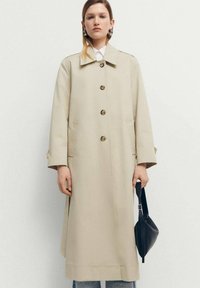 Trench coat bej cu o croială dreaptă, guler mare și nasturi la față. Asortat cu o geantă neagră și blugi bleu deschis.