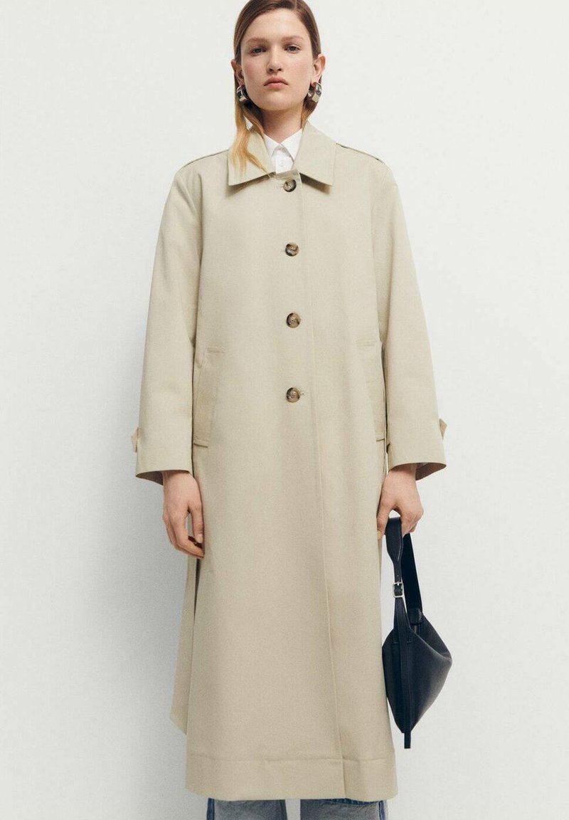 Trench coat bej cu o croială dreaptă, guler mare și nasturi la față. Asortat cu o geantă neagră și blugi bleu deschis.