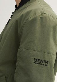 Olivgrön bomberjacka med texturerad yta, svart ribbad krage och broderad "DENIM TOM TAILOR" på ärmen. Dragkedja framtill.