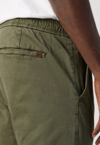 Pantaloni de culoare verde măslin, confecționați dintr-un material ușor, având un brâu elastic, buzunare laterale și o mică etichetă din piele pe spate.