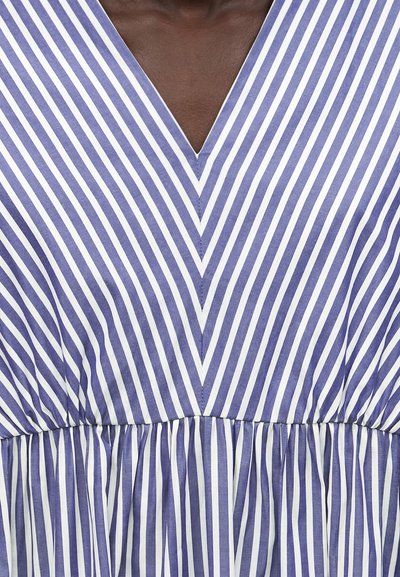 Ρούχο με V-λαιμόκοψη και μπλε και λευκές διαγώνιες ρίγες που συγκλίνουν στο κέντρο, φορεμένο από άτομο με σκούρο χρώμα δέρματος, ύφασμα συγκεντρωμένο στη μέση.