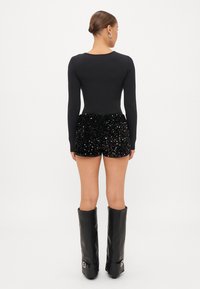 Μαύρο κορμάκι με μακριά μανίκια, συνδυασμένο με μαύρες σορτς με παγιέτες. Το μοντέλο φοράει μαύρες μπότες έως το γόνατο. Απαλή υφή, γυαλιστερή επιφάνεια, εφαρμοστή σιλουέτα.