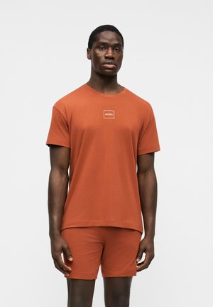Modèle masculin debout, portant un T-shirt à manches courtes couleur rouille avec le logo "HUGO" et un short assorti sur un fond clair uni.