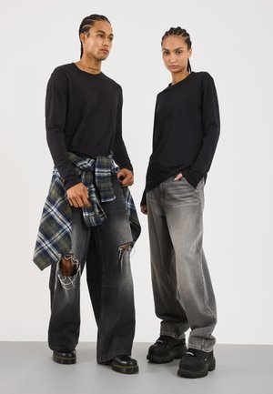 Schwarze Langarmshirts, kombiniert mit weiten grauen Jeans. Eines trägt ein blau-grün kariertes Hemd, das um die Taille gebunden ist. Beide tragen schwarze, klobige Schuhe.