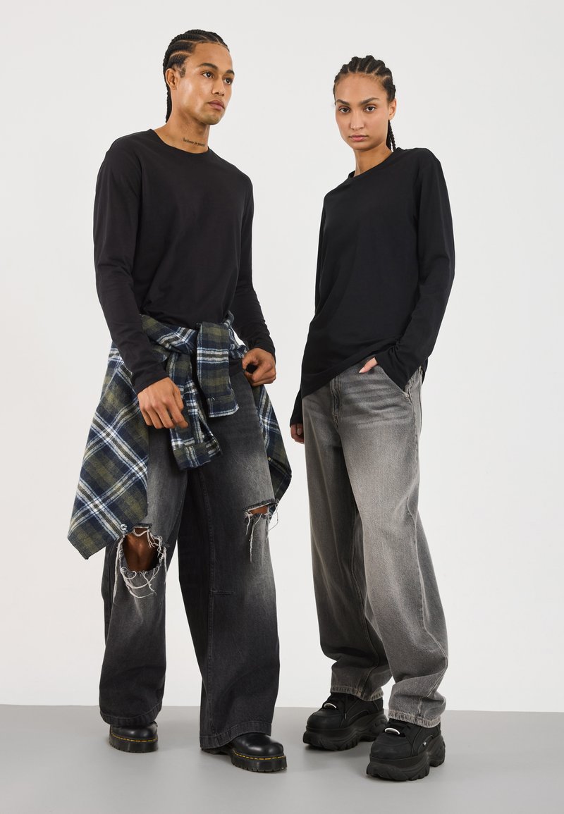 Schwarze Langarmshirts, kombiniert mit weiten grauen Jeans. Eines trägt ein blau-grün kariertes Hemd, das um die Taille gebunden ist. Beide tragen schwarze, klobige Schuhe.