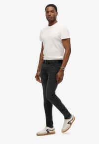 Camiseta blanca de manga corta, jeans negros y zapatillas blancas con detalles en negro. Incluye un cinturón negro y un reloj de pulsera. Pose de pie.