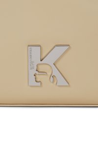Bežna kožna torba s srebrnim "K" logom koji prikazuje siluetu profila i ugravirano ime branda. Glatka tekstura s minimalnim detaljima.