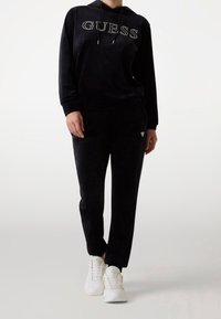 Ensemble sweat à capuche et jogging en velours noir avec logo "GUESS" en strass sur le devant. Associé à des baskets blanches de sport, texture lisse.