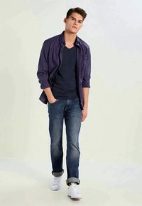 Giovane che cammina con le mani in tasca, indossando una maglietta blu navy, una camicia a quadri, jeans arrotolati e sneaker bianche su uno sfondo semplice.