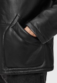 Sort læderjakke med tekstureret finish, sidelomme og løs pasform. Nærbillede af en hånd, der hviler i lommen, med syning detaljer.