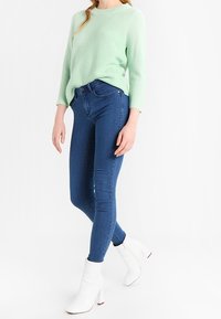 Jasnozielony, oversize sweter o teksturowanym wykończeniu, połączony z niebieskimi wąskimi dżinsami i białymi botkami na obcasie.