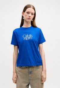 Kuningas sininen puuvillapaita lyhyillä hihoilla, pyöreällä kaula-aukolla ja valkoisella "Calvin Klein" -logolla keskellä rintaa. Rentoa istuvuutta.