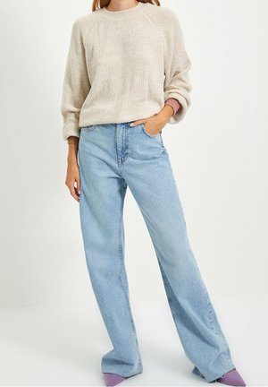 Vrouw draagt een beige gebreide trui, lichtblauwe losse jeans met hoge taille en paarse puntige schoenen, staand met één hand in de zak.