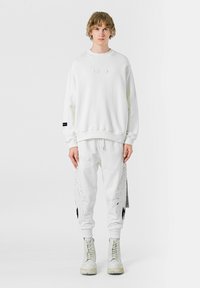 Sudadera blanca de gran tamaño con un panel texturizado, combinada con pantalones deportivos blancos que presentan un diseño de alas y acentos en negro. Botas blancas completan el look.