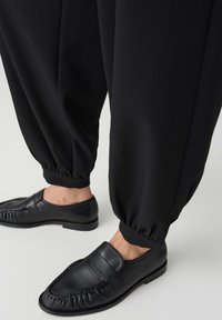 Black leather slip-on shoes worn with black gathered ankle pants on a light gray floor.