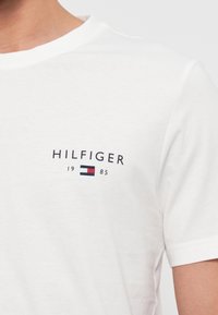 Tricou alb din bumbac, cu textul "HILFIGER" în nuanță de albastru închis, având "1985" și un mic logo dreptunghiular roșu și albastru dedesubt. Guler rotund, mâneci scurte.