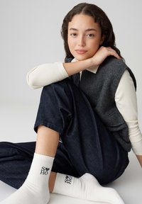 Gilet grigio senza maniche sopra una maglia bianca a maniche lunghe a costine. Jeans a gamba larga blu scuro. Calzini bianchi con testo grafico nero, in posizione seduta.