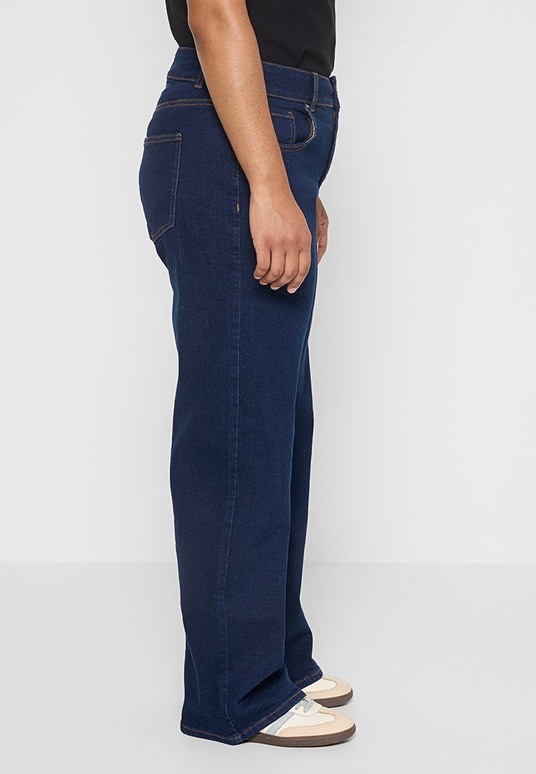 Jean en denim bleu foncé avec une coupe droite, présentant des coutures contrastées et des poches arrière standard. Texture lisse, porté avec des baskets beiges.
