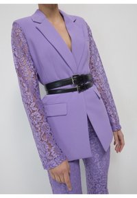 Lawendowa marynarka z koronkowymi rękawami, podkreślona szerokim czarnym paskiem. Charakteryzuje się dopasowanym krojem i klapami z wycięciem, w zestawie z pasującymi koronkowymi spodniami.