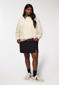 Felpa crema con testo "AMOUR", colletto marrone, abbinata a una gonna nera con uno spacco laterale. Indossata con sneakers crema e calze nere.