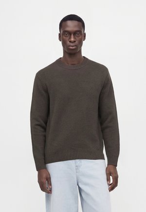 Young man wearing a plain dark brown crewneck sweater and light blue jeans standing against a white background.