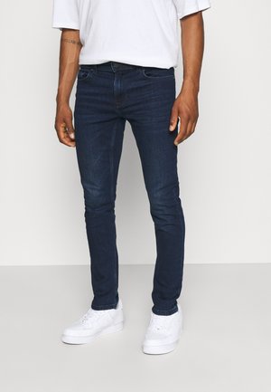 Jean slim bleu foncé en denim, avec une taille mi-haute, un design classique à cinq poches et un léger délavage. Porté avec des baskets blanches.