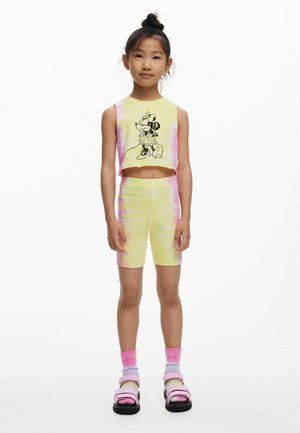 Noor tüdruk kannab kollase ja roosa tie-dye ilma varrukateta crop-topi, millel on Minnie Mouse'i trükk, sobivaid lühikesi pükse, roosasid sokke ja sandaale.