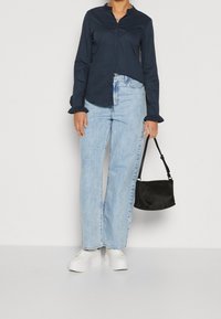 Camisa navy de botones con puños con volantes, combinada con jeans de cintura alta en azul claro, zapatillas de plataforma blancas y un pequeño bolso negro.