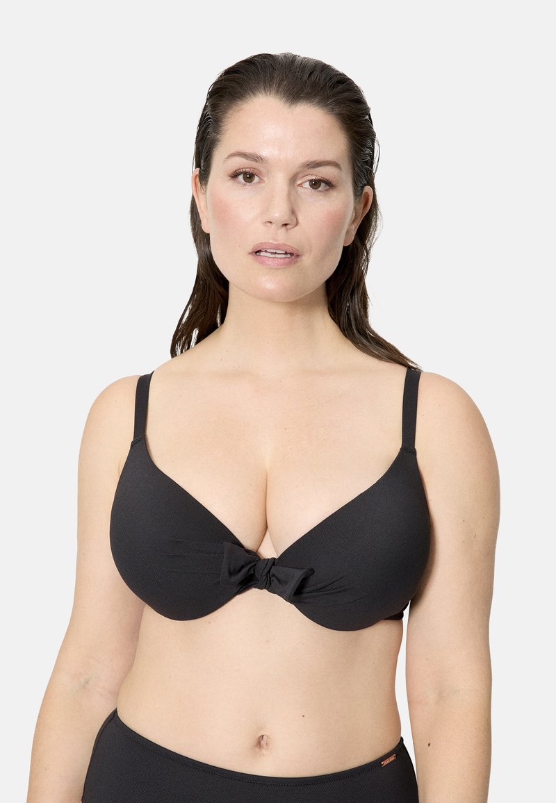 Mujer con el cabello oscuro y mojado, vistiendo un top de bikini negro con un detalle de nudo al frente, sobre un fondo claro y sencillo.
