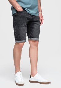 Persona con pantaloncini di jeans neri risvoltati, una camicia blu, scarpe da ginnastica bianche con suola marrone, in piedi con una mano in tasca.