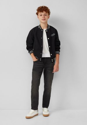 Roodharige jongen staat tegen een grijze muur in een zwarte varsity jack, witte t-shirt, zwarte jeans en witte sneakers met rubberen zolen.
