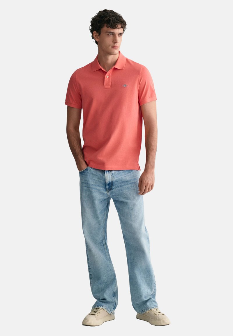 Korallefarbenes Poloshirt mit kurzen Ärmeln, kombiniert mit hellblauen Jeans. Das Outfit wird durch beigefarbene Sneaker ergänzt und zeigt einen lässigen Look.