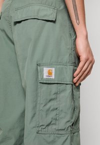 Cargo bukser i et lysegrønt stof med en stor sidelomme og et synligt mærke med et gult logo på en hvid baggrund.