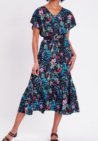 Femme portant une robe midi noire à fleurs avec des motifs roses, bleus et verts, ceinturée à la taille, associée à des talons strap de couleur noire.