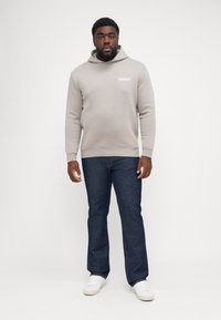 Jasnoszara bluza z kapturem z logo z przodu, w połączeniu z ciemnoniebieskimi dżinsami i białymi trampkami. Bluza ma sznurek ściągający i ściągacze na rękawach.