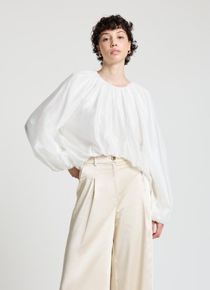 Blouse blanche et ample avec un col froncé et des manches longues, associée à un pantalon plissé taille haute beige clair. Textures lisses.