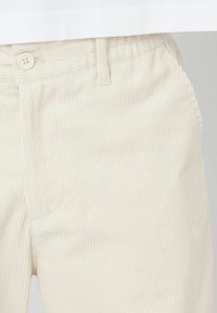 Pantalons en velours côtelé beige avec des côtes verticales, une fermeture à bouton et une poche avant. La taille est cousue et lisse.