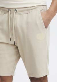 Shorts en polaire beige avec une ceinture élastique, un cordon de serrage, des poches latérales et un logo graphique en forme de cœur sur la jambe gauche.