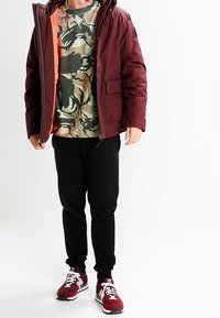 Hombre vistiendo una chaqueta marrón sobre un suéter de camuflaje, joggers negros y zapatillas marrón y blancas de pie contra un fondo blanco.