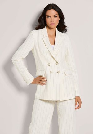Woman wearing a white pinstripe double-breasted blazer and matching wide-leg pants, standing with one hand on her hip.