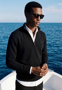 Jeune homme portant des lunettes de soleil et un pull noir zippé par-dessus une chemise blanche, debout sur un bateau avec une mer bleue calme en arrière-plan.